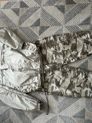 Beige camo vindjacka med huva - Snygg vindjacka i beige och grått camomönster. Jackan har dragkedja framtill, justerbar midja med snöre och elastiska muddar vid ärmarna. Materialet är tunt och vindtätt, perfekt för blåsiga dagar. Stora fickor på sidorna och huva för extra skydd.