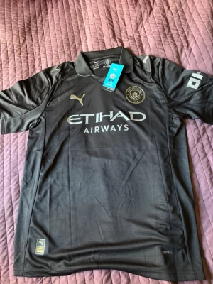 Manchester City bortatröja Haaland - Snygg svart Manchester City bortatröja från Puma med guldiga detaljer och Etihad Airways-tryck på bröstet. Nummer 9 och HAALAND på ryggen. Tillverkad i lätt och ventilerande material, perfekt för fotboll eller träning.