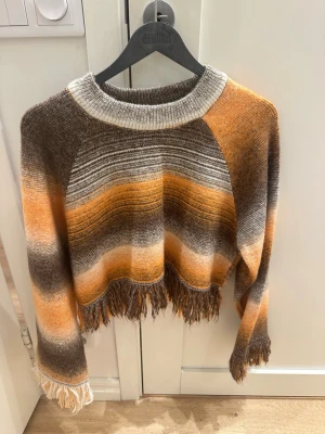 Randig cropad stickad tröja Zara - Säljer en stickad cropad tröja från Zara med breda ränder i brunt, orange, beige och vitt. Tröjan har fransad nederkant och muddar vid ärmslut. Mjuk och skön med avslappnad passform, perfekt för höst och vinter.