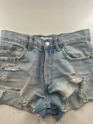 Ljusblåa högmidjade jeansshorts  - Säljer ett par ljusblåa high waist jeansshorts med slitna detaljer och råa kanter. Shortsen har fyra fickor, bälteshällor och knappgylf. Perfekta för en avslappnad och trendig look.