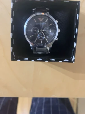 Emporio Armani silverfärgad klocka - Stilren klocka från Emporio Armani i silverfärgat rostfritt stål. Urtavlan är svart med flera små urtavlor och tydliga indexmarkeringar. Klassisk länkarmband och diskret logga på både urtavla och baksida. Perfekt accessoar för en trendig look.