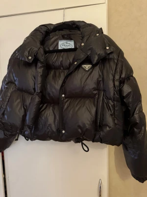 Svart dunväst från Prada - Snygg svart dunväst från Prada med avtagbar huva och ikonisk trekantslogga på bröstet. Västen har en glansig finish, stora fickor och dragsko i nederkant för extra stilpoäng. Perfekt för dig som vill ha en trendig och varm look. Ej äkta