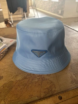 Ljusblå bucket hat från Prada pris går diskutera  - Snygg ljusblå bucket hat från Prada i glansigt nylonmaterial. Klassisk form med bred, nedåtlutande brätte och ikoniska metalltriangeln med logga framtill. Perfekt accessoar för en trendig och avslappnad stil. 