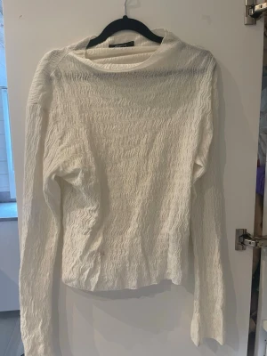 Vit långärmad topp med vågigt mönster - Säljer en vit långärmad topp med vågigt strukturerat mönster. Toppen har en något lös passform och är tillverkad i ett tunt, lätt material som känns luftigt. Perfekt att styla med jeans eller kjol för en trendig look.