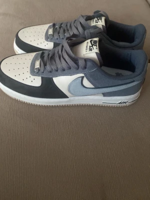 Nike Air Force 1 låga sneakers grå/blå - Nike Air Force 1 sneakers i lågt utförande med vit tåbox, gråblå mockadetaljer och klassisk Swoosh i ljusblått. Skorna har perforerad tå, vita sulor och grå snören. Perfekt för dig som gillar stilrena och tidlösa sneakers.