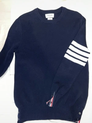 Marinblå stickad tröja Thom Browne - Snygg marinblå stickad tröja från Thom Browne med rund halsringning och vita ränder på ena ärmen. Tröjan har ribbade muddar och knappar vid nederkanten samt en detalj med röd, vit och blå rand. Tillverkad i Japan.