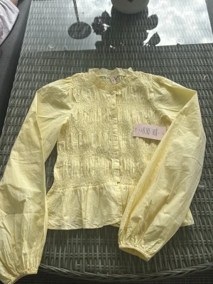 Gul blus från Nelly - Söt ljusgul blus från Nelly med smockad front, volangkant vid halsen och knappar framtill. Blusen har långa ärmar med resår vid ärmslut och lätt rynkad nederkant. Perfekt för dig som vill ha en feminin och trendig look. Helt oanvänd 