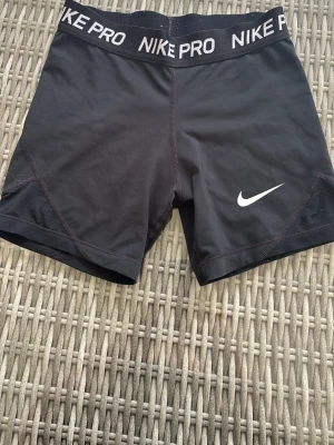 Svarta Nike Pro träningsshorts - Säljer ett par svarta Nike Pro träningsshorts med vit logga på benet och bred resår i midjan med texten 'NIKE PRO'.  Lappen är avklippt då den kliade. 