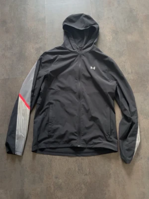 Svart vindjacka från Under Armour - Snygg svart vindjacka från Under Armour med huva och hel dragkedja. Jackan har grå och vita detaljer på ärmarna samt en röd linje för extra stil. Liten logga på bröstet och praktiska fickor framtill. Perfekt för blåsiga dagar och sportiga outfits.   Passar både S och M