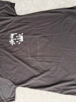 Svart Stüssy t-shirt med krona - Svart t-shirt från Stüssy med stort vitt tryck av krona och logga på ryggen samt mindre tryck på bröstet. Klassisk passform och rund hals. Perfekt för dig som gillar streetwear och vill sticka ut med en ikonisk design.