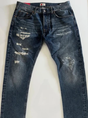 Blå jeans från Dondup - Säljer ett par blå jeans från Dondup med snygga slitningar och distressed detaljer på både fram- och baksida. Klassisk femficksmodell med raka ben och knappgylf. Jeansen är gjorda i denim och har en cool vintagekänsla.