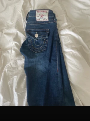 Blå True Religion jeans med kontrastsömmar - Säljer ett par klassiska blå jeans från True Religion med tydliga kontrastsömmar och ikonisk fickdesign bak. Jeansen har normal midja, raka ben och silverfärgade knappar. Materialet är slitstark denim i bomull. Perfekta för en avslappnad och trendig look.