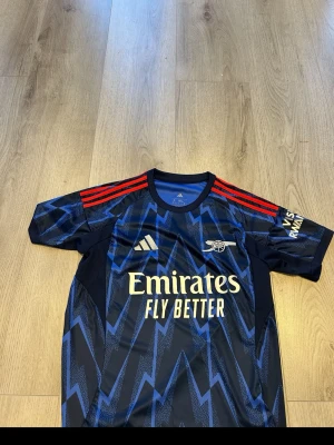 Arsenal x Adidas blå matchtröja - Snygg Arsenal matchtröja i mörkblått med blixtmönster i ljusblått och röda ränder på axlarna. Adidas-logga och Arsenal-emblem på bröstet samt Emirates Fly Better-tryck framtill. Tillverkad i lätt och ventilerande syntetmaterial, perfekt för träning eller match.