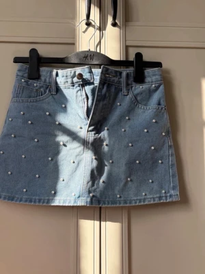 Blå jeanskjol med pärldetaljer H&M - Snygg blå jeanskjol från H&M med pärldetaljer över hela kjolen. Klassisk fickmodell med knapp och dragkedja framtill. Perfekt för dig som vill ha en trendig och ungdomlig look.