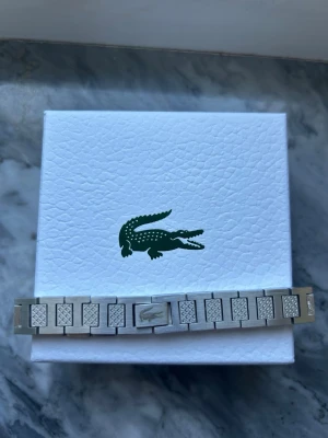 Silverfärgat armband från Lacoste - Snyggt armband i silverfärg från Lacoste med rektangulära länkar och diskreta mönstrade detaljer på vissa länkar. Armbandet har en stilren och modern design med Lacoste-loggan på låset. Perfekt accessoar för dig som gillar minimalistisk stil.