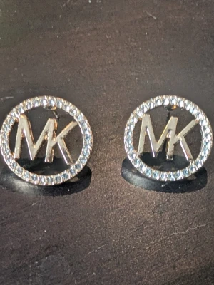 Guldtonade MK örhängen med stenar - Snygga örhängen i guldtonad metall. De är runda med MK-loggan i mitten och har en ram av gnistrande stenar som ger extra bling. Perfekta för dig som gillar stilrena accessoarer med lite lyxig känsla.