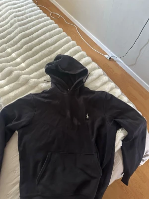 Svart hoodie från Polo Ralph Lauren - Snygg svart hoodie från Polo Ralph Lauren med klassisk huva och dragsko. Ikoniska Polo-loggan broderad på bröstet. Hoodien har en stor magficka och är tillverkad i mjukt material som känns skönt mot huden. Perfekt för en avslappnad och stilren look.