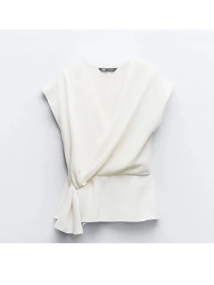 Vit omlottopp från Zara - Säljer en stilren vit omlottopp från Zara med knyt i sidan och v-ringad hals. Toppen har korta ärmar och en lätt figurnära passform som ger en snygg siluett. Perfekt att matcha med jeans eller kjol för en trendig look.