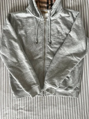 Burberry Zip Hoodie – Grå 🤍 - Snygg och bekväm Burberry hoodie med dragkedja i gråmelerad färg. Klassisk Burberry-rutig detalj på insidan av huvan och broderad logotyp på bröstet. Perfekt för både vardag och en mer uppklädd casual stil.  ✔️ Jättebra skick ✔️ Mjuk och skön passform ✔️ Dragkedja och huva med snörning  Säljes eftersom den inte kommer till användning. Hör gärna av dig vid frågor eller för fler bilder!
