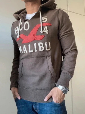 Brun hoodie från Hollister Malibu - Mörkbrun hoodie från Hollister med stor röd fågel och texten 'HCO 54 MALIBU' broderat framtill. Klassisk känguruficka, vita dragsnören i huvan och ribbade muddar. Perfekt för en avslappnad och cool stil.