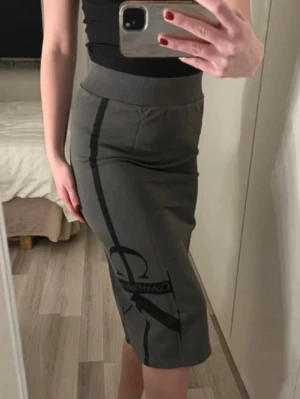 Grå kjol från Calvin Klein Jeans - Säljer en grå, figurnära kjol från Calvin Klein Jeans med svart logga och vertikal linje framtill. Kjolen har bred resår i midjan och går ner till knäna. 