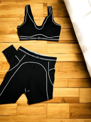 Svart träningsset från Alo - Snyggt svart träningsset från Alo med sport-bh och tights. Setet har vita kontrastsömmar och en sportig design. Perfekt för gymmet eller yoga. Materialet är stretchigt och följsamt för maximal komfort.
