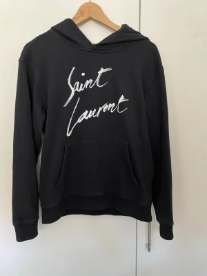 Svart hoodie från Saint Laurent - Cool svart hoodie från Saint Laurent med vit handskriven logga på bröstet. Klassisk huva och stor känguruficka framtill. Mjukt material och ribbade muddar vid ärmslut och nederkant. Perfekt för en avslappnad och trendig look.