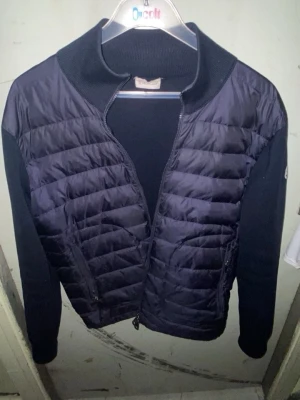 Mörkblå cardiganjacka från Moncler - Snygg äkta mörkblå cardiganjacka från Moncler med quiltad front och stickade ärmar. Jackan har dragkedja framtill, hög krage och Moncler-logga på ärmen. Perfekt för lager på lager och en clean streetstyle-look.