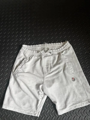 Vita shorts från Moncler med snörning - Säljer ett par vita shorts från Moncler med snörning i midjan och två sidofickor. Shortsen har Moncler-logga broderad på vänster ben och är tillverkade i mjuk bomull, perfekta för sommardagar.