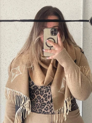 Beige Vuitton halsduk med fransar - Snygg beige halsduk från  Vuitton med ikoniska monogram och blommönster i vitt, brunt, svart och gult. Halsduken har långa fransar i ändarna och är tillverkad i ett mjukt, vävt material som känns lyxigt och trendigt.