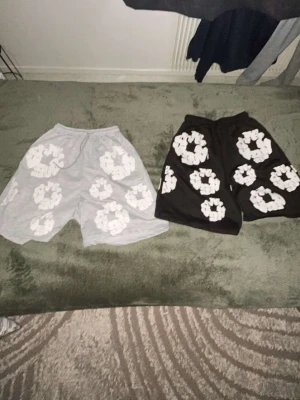 Två par shorts Denim Tears - Säljer två par shorts, ett par i ljusgrått och ett par i svart, båda med stora vita blommiga mönster. De har en loose passform och elastisk midja för skön komfort. Perfekta för sommarens alla dagar.