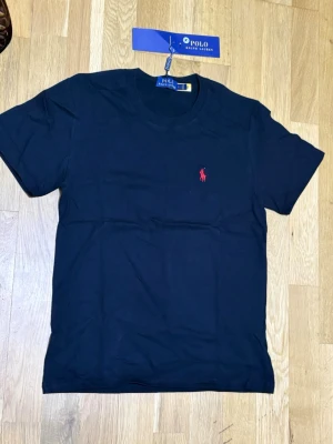 Svart t-shirt från Polo Ralph Lauren - Klassisk svart t-shirt från Polo Ralph Lauren med röd broderad logga på bröstet. T-shirten har rund halsringning och korta ärmar. Tillverkad i mjuk bomull för en bekväm känsla och stilren look.