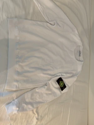 Vit sweatshirt från Stone Island - Vit sweatshirt från Stone Island med klassisk rund hals och ikonisk svart patch med logga på vänster ärm. Tröjan har ribbade muddar vid ärmslut och nederkant, perfekt för en clean och stilren look.