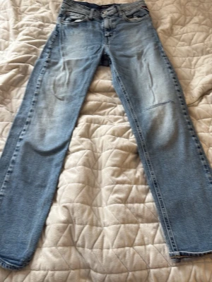Ljusblå raka jeans replay med lagning - Säljer ett par ljusblå jeans med rak passform och klassisk femficksdesign. Jeansen har en tydlig lagning på ena benet och slitningar framtill för en avslappnad look. Materialet är denim i bomull. Perfekta för dig som gillar vintagekänsla.