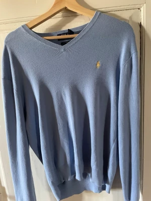 Ljusblå tröja från Ralph Lauren - Ljusblå långärmad tröja från Ralph Lauren med klassisk v-ringning och den ikoniska broderade loggan i gult på bröstet. Tröjan har ribbade muddar vid ärmslut och nederkant. Perfekt för en stilren och avslappnad look.