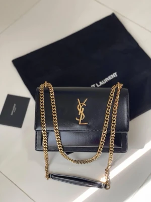 Svart axelväska från Saint Laurent - Snygg svart axelväska från Saint Laurent i slätt läder med ikoniskt YSL-emblem i guld framtill. Väskan har en elegant guldfärgad kedjerem och stilren, fyrkantig form. Perfekt för dig som vill ha en lyxig och tidlös accessoar.