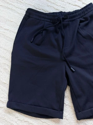 Mörkblå shorts med snörning - Mörkblå shorts med snörning i midjan och uppvikta benslut. De har en avslappnad passform och är tillverkade i ett mjukt material som passar perfekt för varma dagar. Shortsen har fickor på sidorna och elastisk midja för extra komfort. Nypris 329kr
