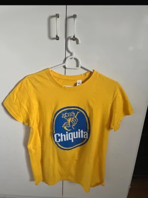 Gul Chiquita t-shirt i bomull - Gul t-shirt med Chiquita-logga i blått och vitt framtill. Klassisk rund halsringning och korta ärmar. Tillverkad i mjuk bomull, perfekt för en avslappnad och färgglad stil.