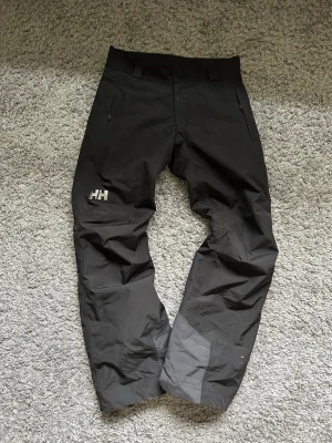 Svarta täckbyxor från Helly Hansen - Svarta täckbyxor från Helly Hansen med HH-logga på låret. Byxorna har raka ben, förstärkta partier vid knä och benslut samt två dragkedjeförsedda fickor. Materialet är slitstarkt och vattenavvisande, perfekt för kalla dagar utomhus.
