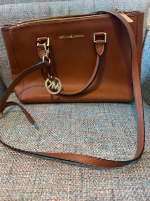 Brun andväska från Michael Kors - Snygg brun handväska från Michael Kors i slätt läder med guldfärgade detaljer och logotyp. Väskan har både axelrem och handtag samt en dekorativ MK-berlock. Perfekt för dig som vill ha en klassisk och stilren accessoar.