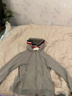 Grå Moncler hoodie med dragkedja - Snygg grå hoodie från Moncler med dragkedja framtill och två fickor. Huvan har ett coolt innerfoder med röd, vit och marinblå rand. Tillverkad i mjukt bomullsmaterial som känns skönt mot huden. Perfekt för en avslappnad och trendig look.DEN ÄR I STORLEK M MEN PASSAR SOM xs