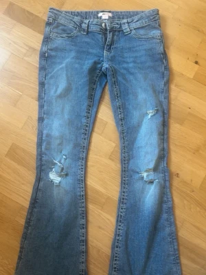 Blå bootcut jeans med slitningar - Snygga blå bootcut jeans med slitningar på båda benen och broderade bakfickor. Jeansen har klassisk femficksdesign och är tillverkade i denim. Perfekta för dig som gillar en avslappnad och trendig stil.