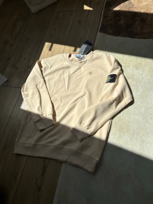 Beige stickad tröja från Stone Island - Snygg beige stickad tröja från Stone Island med klassisk rund halsringning och ribbade muddar. Tröjan har den ikoniska svarta logotypen med gul brodyr på vänster ärm. Perfekt för en clean och stilren look.