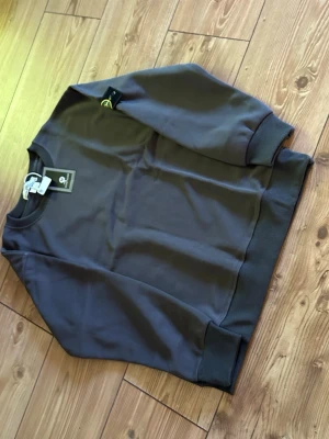 Mörkgrå sweatshirt från Stone Island - Mörkgrå sweatshirt från Stone Island med klassisk rund hals och ribbade muddar. Tröjan har den ikoniska Stone Island-loggan på vänster ärm och är tillverkad i mjukt material som känns skönt mot huden. Perfekt för dig som gillar stilrena och trendiga plagg.