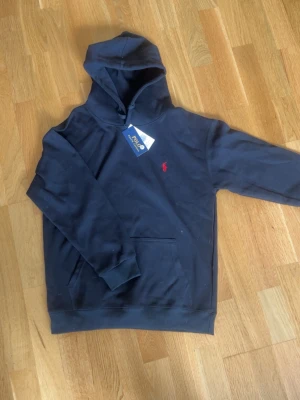 Svart hoodie från Polo Ralph Lauren - Snygg marinblå hoodie från Polo Ralph Lauren med klassisk huva och dragsko. Ikoniska röda loggan broderad på bröstet och stor ficka framtill. Tillverkad i mjuk bomull för skön känsla. Perfekt för en avslappnad och stilren look.
