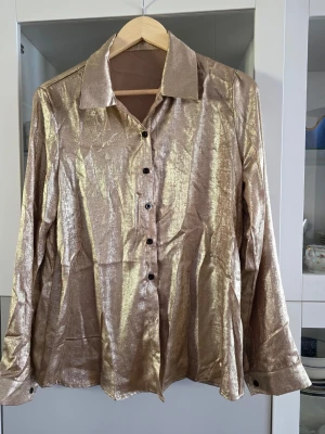 Glittrig guldskjorta med knappar - Säljer en glittrig skjorta i guld med klassisk krage och svarta knappar framtill. Skjortan har lång ärm och ett riktigt statement-plagg för dig som vill sticka ut. Perfekt att styla med jeans eller kjol för en trendig look. Använd en gång på fest, perfekt om du ska på fest med tema 