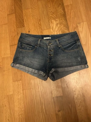 Blå lowrise jeansshorts med knappar 🫐 - Säljer ett par jättefina lågmidjade jeansshorts från Terranova, köpta second hand i mycket bra skick. Shortsen har en klassisk femficksdesign, knappgylf och kontrastsömmar för en snygg detalj. Perfekta till sommaren och lätta att matcha med olika stilar! 💗