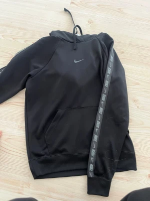 Svart Nike hoodie med logga - Snygg svart hoodie från Nike med klassisk swoosh-logga på bröstet och Nike-band längs ärmarna. Hoodien har huva med dragsko, känguruficka framtill och är tillverkad i ett mjukt, sportigt material med meshfoder i huvan. Perfekt för en avslappnad streetwear-look.