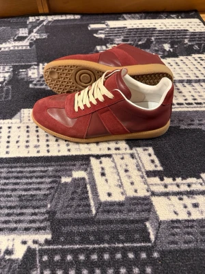 Vinröda sneakers med mockadetaljer - Säljer ett par vinröda sneakers med snygga mockadetaljer och vita skosnören. Skorna har en klassisk låg silhuett och gummisula i beige. Insidan är vit och ovandelen kombinerar både mocka och slätt läder för en stilren look.