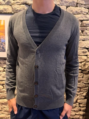 Mörkgrå cardigan med knappar - Stilren mörkgrå cardigan från peir one med v-ringning och fem knappar framtill. Tröjan har långa ärmar, ribbade muddar och nederkant. Perfekt att slänga över en t-shirt för en clean look. Materialet känns mjukt och lätt, passar året runt.